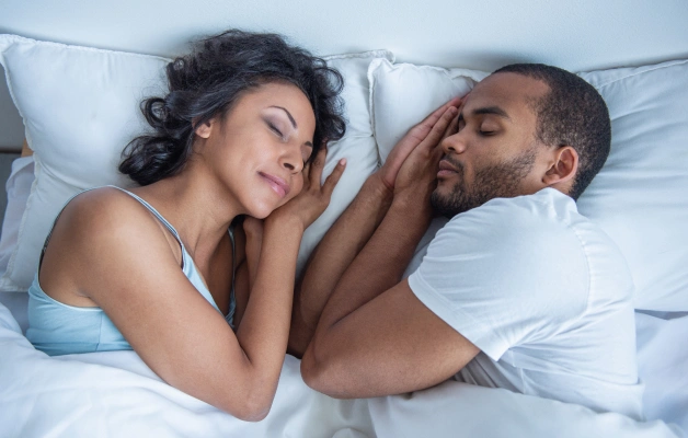 Couple lying in bed together on white sheets, appearing thoughtful or concerned about sleep issues.